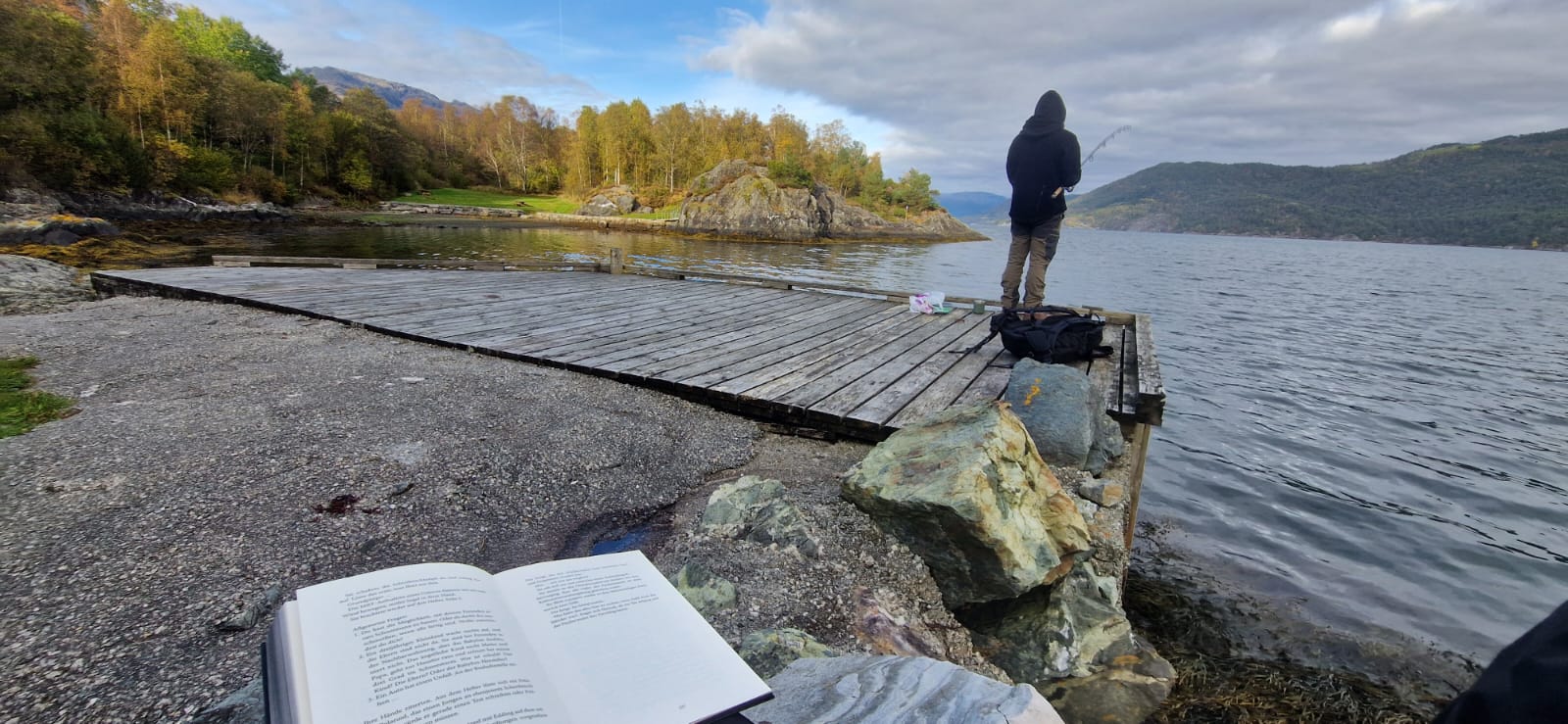 Person sitzt auf einem Holzsteg, liest ein Buch und angelt in einem ruhigen Fjord, von hinten fotografiert.